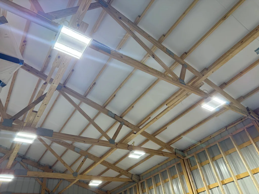 White sprayed foam across a pole barn gable ceiling with timber purlins and strip lights