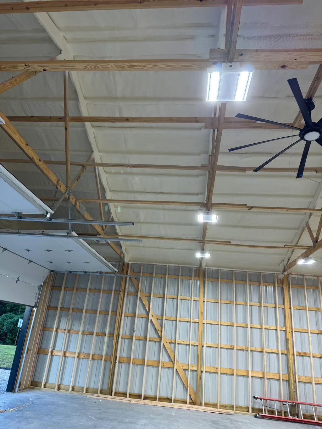 Large dark ceiling fan under a foamed barn ceiling, garage door open to green trees