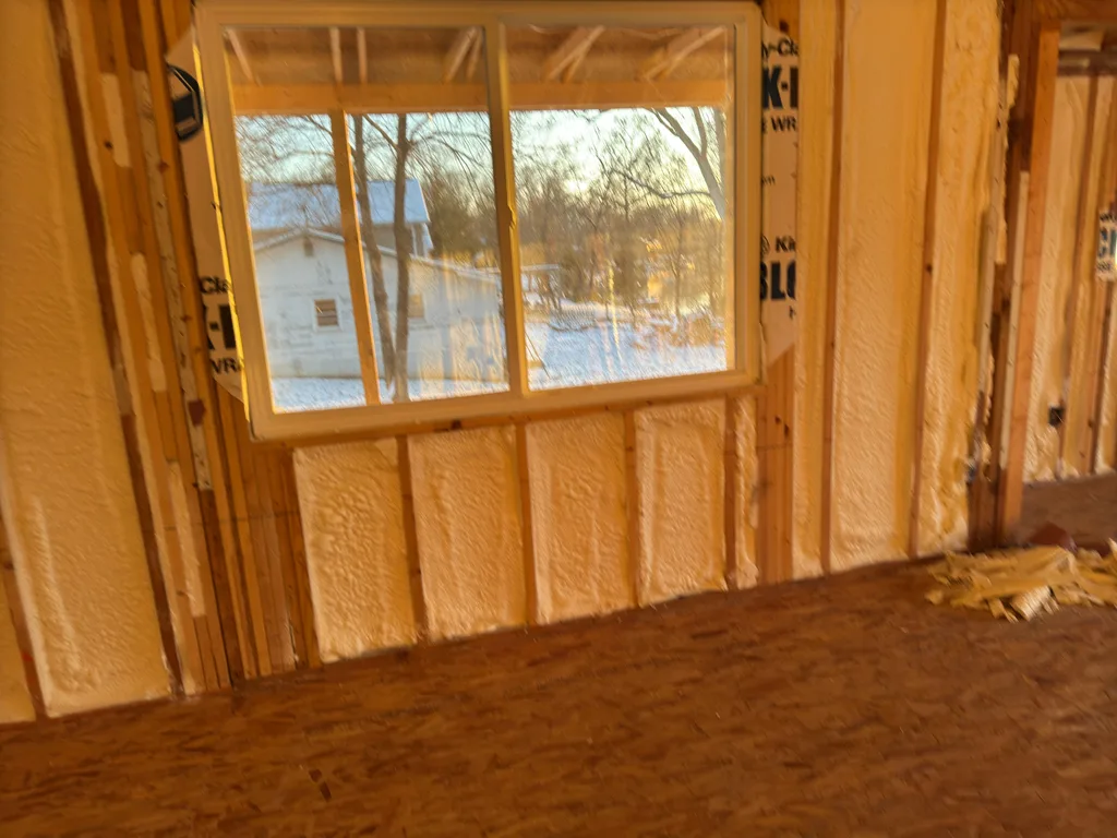 Foamed stud wall around a window, low sun casting orange light across the plywood floor