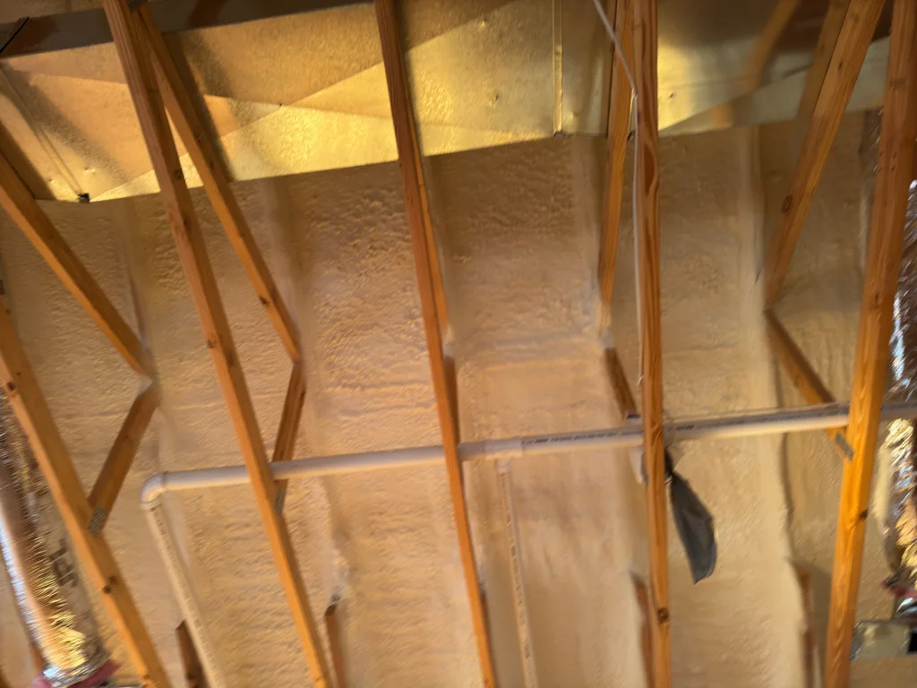 Foamed ceiling and rafters under warm light with a duct running along the wall